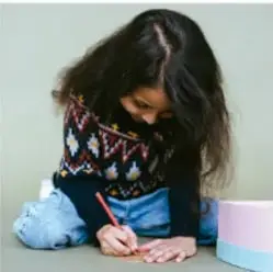 Niña sentada escribiendo en un cuaderno con lápiz, cabello largo, suéter de colores, fondo claro.
