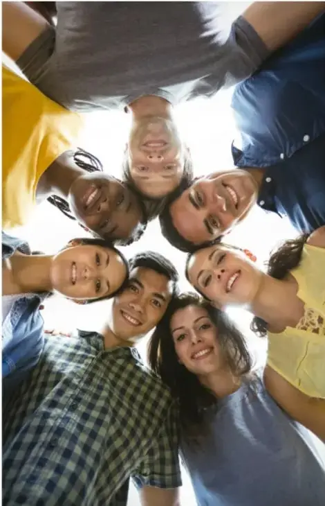 Fotografía tomada desde abajo (contrapicado) de un grupo diverso de jóvenes sonrientes que miran hacia la cámara, rodeando el encuadre, representando diversidad y comunidad.