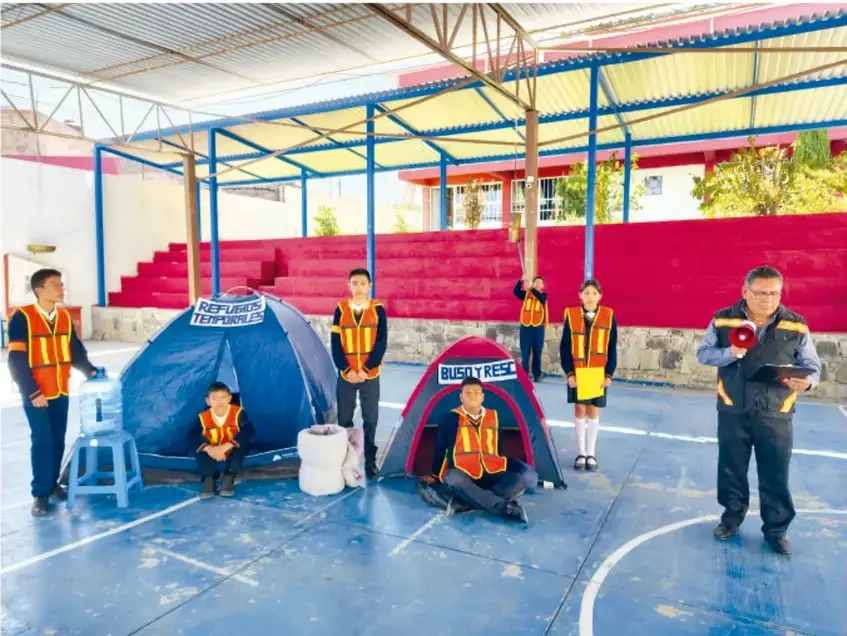 Fotografía de estudiantes de telesecundaria con chalecos naranjas de simulacro en una cancha techada; hay una carpa azul rotulada 'REFUGIOS TEMPORALES', una carpa roja 'BUS & RESCUE', un rescatista adulto con megáfono y alumnos con petos de seguridad.