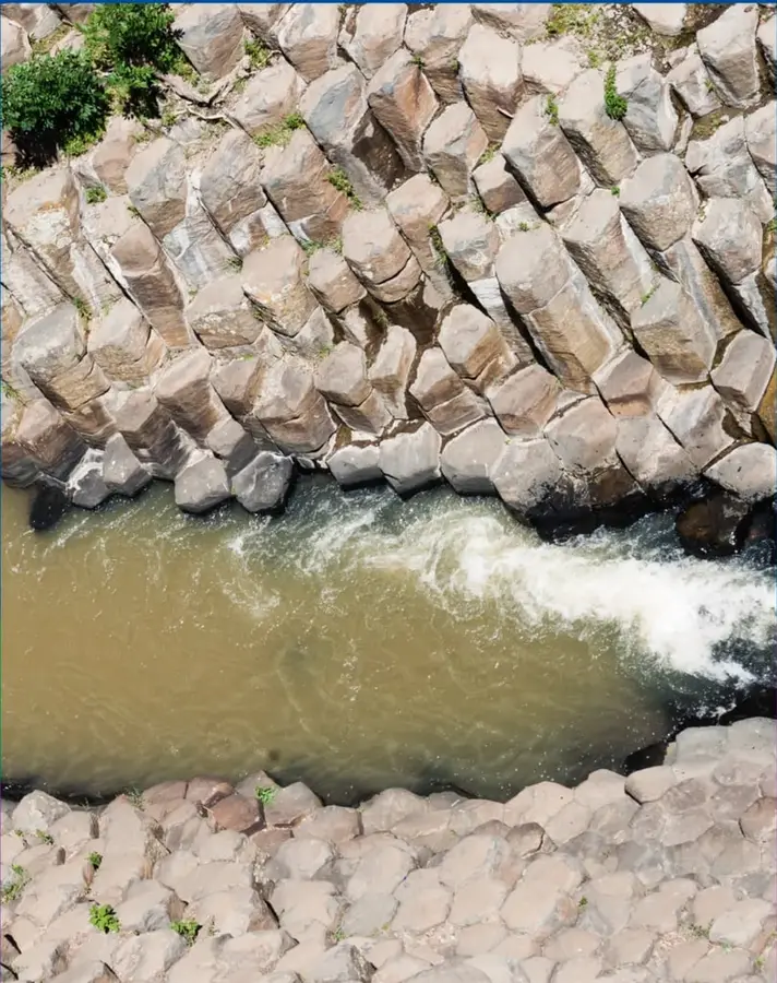 Fotografía aérea de columnas de basalto de forma poligonal (hexagonal) formando un escarpe rocoso, con un río de agua turbia y espuma blanca fluyendo al pie de las columnas; vegetación pequeña creciendo en las juntas de la roca.