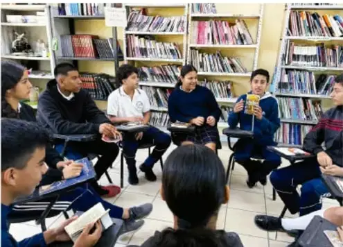 Fotografía de un grupo de estudiantes de telesecundaria sentados en círculo en una biblioteca escolar, leyendo y comentando un folleto.