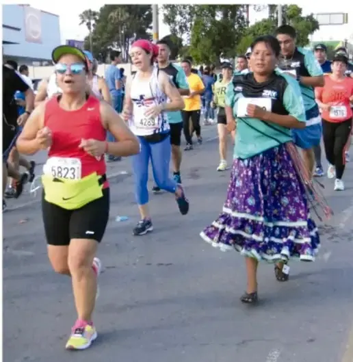 Fotografía de un maratón donde participan corredores con ropa deportiva convencional junto a una mujer indígena que corre con la vestimenta típica de su comunidad (falda larga de color, rebozo), ilustrando la tradición del pueblo ralámuli.