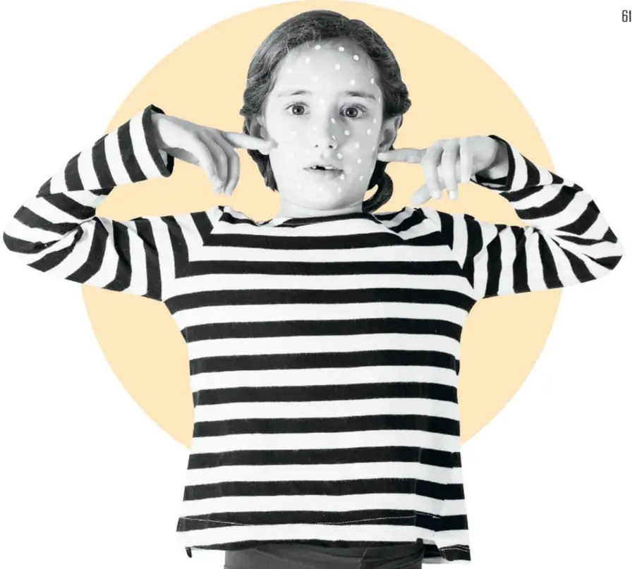 Fotografía en blanco y negro de una niña con camiseta de rayas negras y blancas, con puntos blancos pintados en el rostro, señalándose las mejillas con los índices; el fondo tiene un círculo color durazno. Ilustra la expresión facial y corporal propia de la mímica.