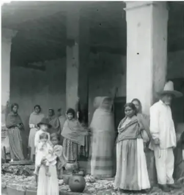 Fotografía en blanco y negro de un grupo de personas, entre ellas mujeres con rebozo y un hombre con sombrero, reunidas junto a columnas de piedra de un edificio antiguo.