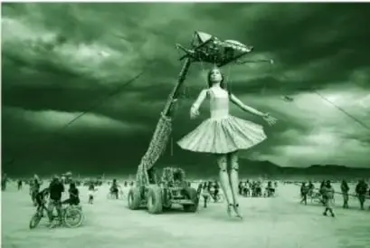 Fotografía (en tinta verde del libro) del festival Burning Man: marioneta gigante de bailarina de ballet con tutú rosa suspendida de una grúa en un desierto, con personas alrededor, algunas en bicicleta y otras sujetando cuerdas, bajo un cielo nublado.