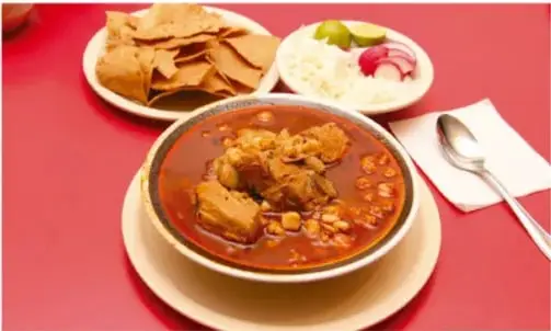 Platillo hondo con caldo rojo y piezas de carne (tipo pozole o guiso), acompañado de totopos, cebolla, rábano y limón sobre mantel rojo.