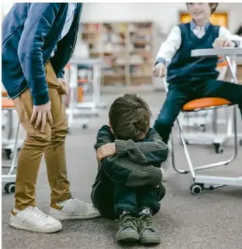 Fotografía de un niño sentado en el suelo con las rodillas abrazadas y la cabeza baja, en actitud de tristeza o miedo. Detrás, un adulto de pie y otro niño sentado en una silla, en un ambiente que sugiere una escuela o aula.