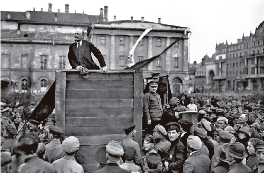 Fotografía en blanco y negro de un líder (Vladímir Lenin) de pie sobre una tribuna de madera, dirigiéndose a una multitud en una plaza pública. Al fondo se observan edificios y una bandera. La escena corresponde a un discurso durante la Revolución Rusa.