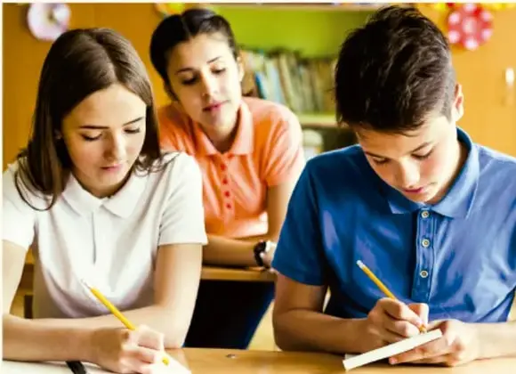 Fotografía de tres estudiantes trabajando en equipo: dos escriben en sus cuadernos y una compañera los observa.