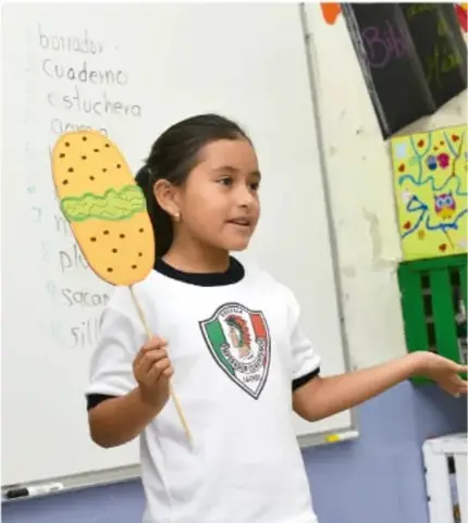 Niña sosteniendo una figura de cartón con forma de taco, frente a un pizarrón con palabras en inglés y dibujos.