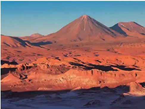 Paisaje del desierto de Atacama: cerros y arena de color rojizo, cielo despejado y ausencia de vegetación; en la base se observa una superficie clara, posiblemente salar o nieve.