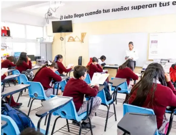 Fotografía de un aula de telesecundaria: estudiantes con uniforme rojo sentados en pupitres azules frente a una maestra que explica junto a la pizarra; en la pared se lee el lema «El éxito llega cuando tus sueños superan tus e…».