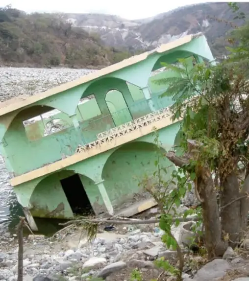 Fotografía de una construcción de concreto pintada de verde, inclinada y dañada, rodeada de escombros y vegetación al pie de un cerro, ilustrando los efectos de un desastre natural sobre el relieve.