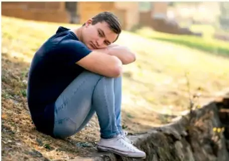 Fotografía de un joven sentado al aire libre, con los brazos abrazando las rodillas y la cabeza apoyada en ellas, con expresión melancólica o reflexiva, sobre un fondo verde desenfocado.