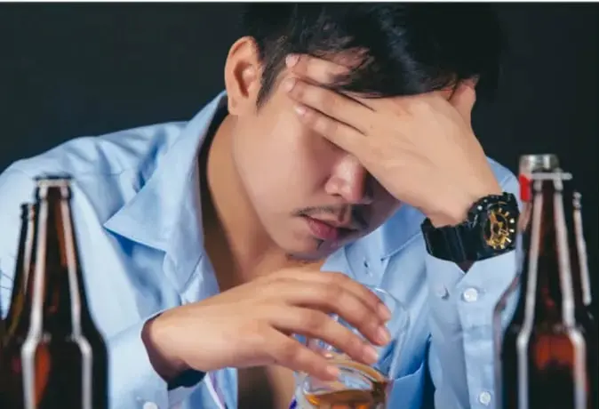 Foto de un joven con botellas de alcohol, con la mano en la frente, aparentando malestar o dolor de cabeza.