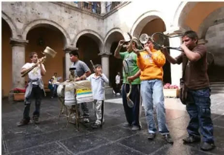 Grupo de personas (adultos y niños) tocando instrumentos de percusión en un patio o plaza colonial, en actividad comunitaria.