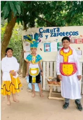 Tres estudiantes con disfraces y artesanías frente a un mural que dice 'Cultura Chichimecas y Zapotecos', presentando contenido cultural de manera comunitaria.