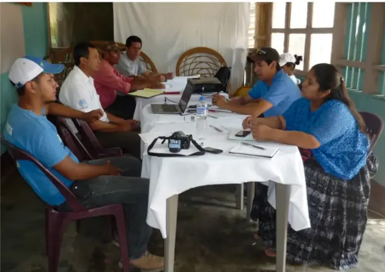 Fotografía de una asamblea escolar: varias personas sentadas alrededor de una mesa con mantel blanco, computadora y documentos, en un salón con ventanas.