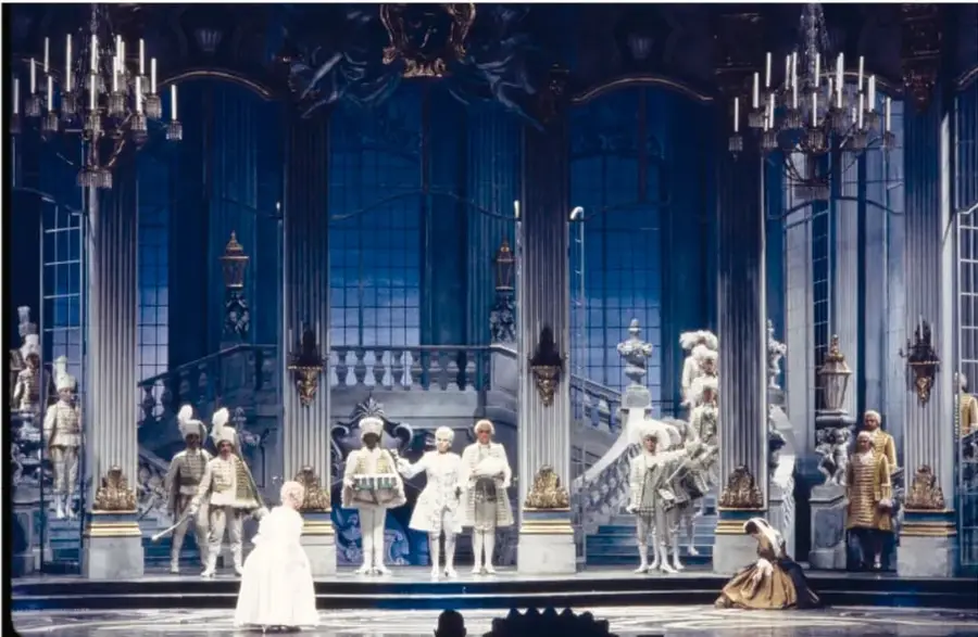 Fotografía a color de una escenografía teatral de ópera con columnas, escalinatas y arañas; actores con vestuario del siglo XVIII en escena.
