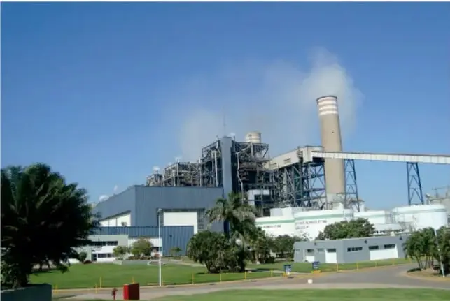 Fotografía de una planta termoeléctrica con chimenea alta que emite humo, rodeada de vegetación y áreas verdes.