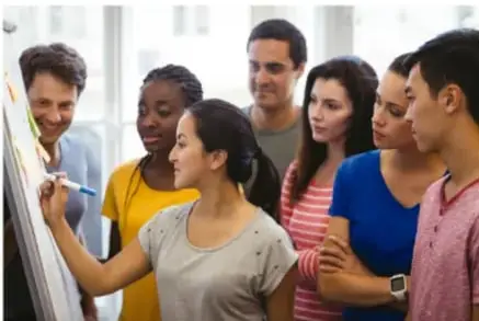 Fotografía de un grupo diverso de jóvenes y adultos de distintos ethnicidades observando y participando en una sesión de trabajo frente a un rotafolio, ilustrando la inclusión social.