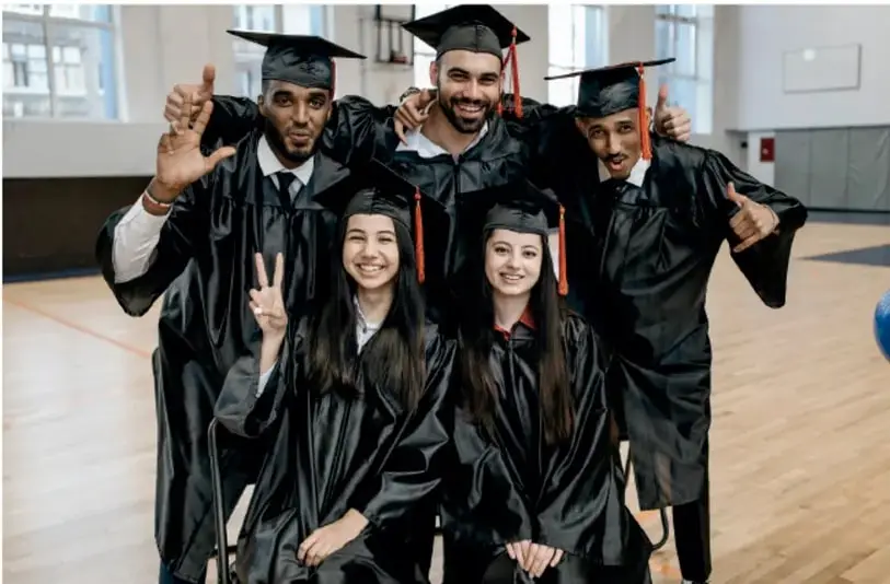 Fotografía de cinco jóvenes con toga y birrete de graduación, sonriendo y haciendo señas de aprobación en un gimnasio o patio escolar.