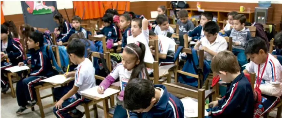Fotografía de un aula de primaria con estudiantes sentados en sus pupitres, trabajando en equipo con cuadernos y lápices.