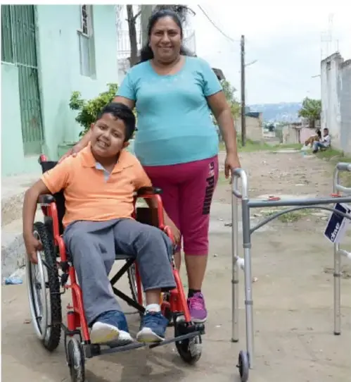 Fotografía de una mujer adulta de pie junto a un joven en silla de ruedas, en un camino de tierra con casas al fondo; ambos sonríen y la mujer apoya la mano en el hombro del joven. Representa la inclusión y el acompañamiento.