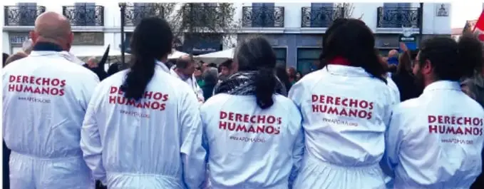 Fotografía de un grupo de personas de espaldas vistiendo chambros blancos con la leyenda 'DERECHOS HUMANOS' impresos en rojo, en una marcha o movilización pública.