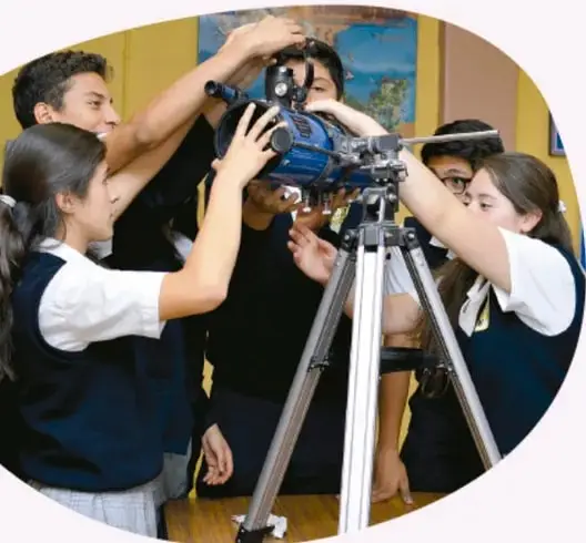 Grupo de estudiantes de primaria observando el cielo con un telescopio montado en trípode, guiados por una maestra.