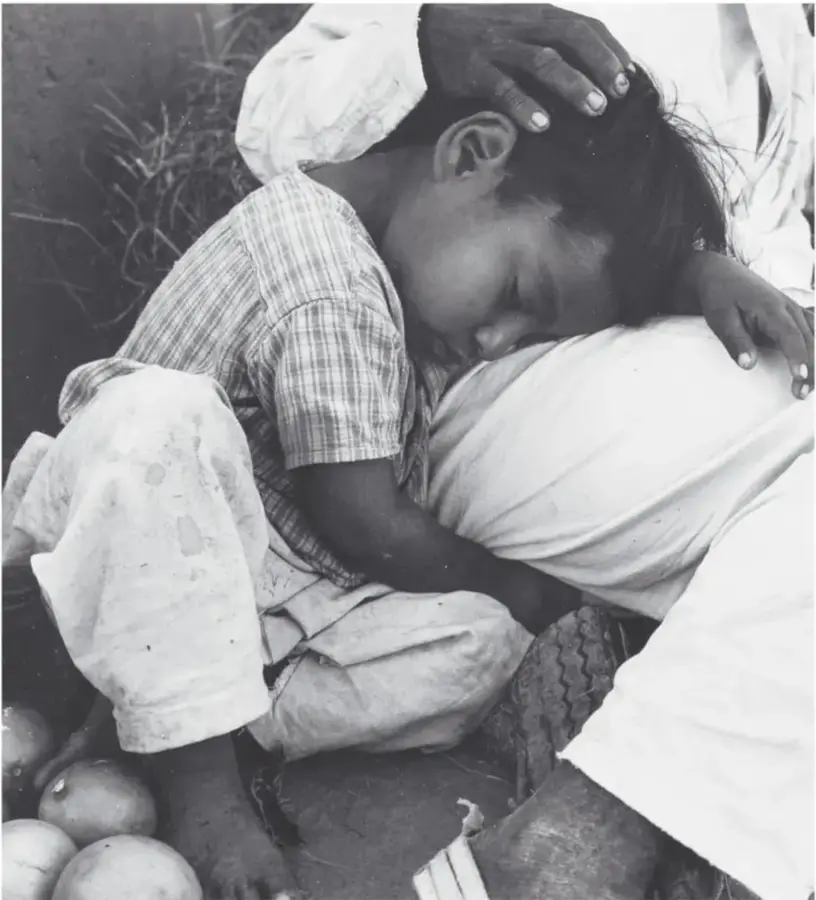 Fotografía en blanco y negro de una niña dormida sobre una persona adulta sentada, junto a naranjas en el suelo de un puesto; un pie descalzo y una sandalia se ven al frente. Puesto de naranjas, 1985, Axochiapan, Morelos, de Mariana Yampolsky (1925-2002).