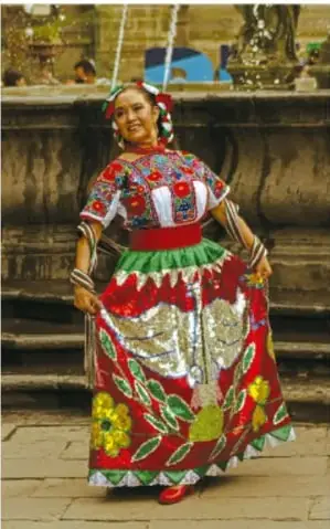 Fotografía de una mujer con traje típico mexicano bordado en rojo, verde y blanco, con flores en la falda, de pie frente a una fuente de piedra; representa la riqueza del vestuario tradicional de una región.