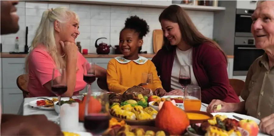 Fotografía de una familia multigeneracional reunida en torno a la mesa para Thanksgiving: abuela rubia, niña afrodescendiente, mujer, abuelo, todos sonriendo con copas de vino, pavo asado, maíz y otros platillos.