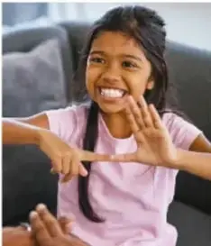Niña sonriente con playera rosa que señala con ambas manos hacia un lado, en un ambiente escolar.