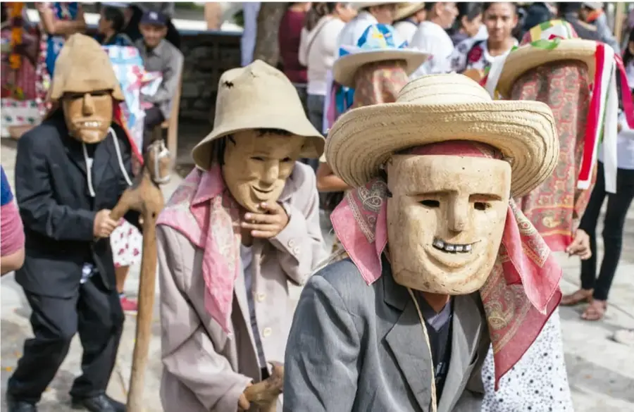 Danzante con máscara de viejito sonriente, saco oscuro y sombrero de palma, frente a una puerta de madera tallada con motivos florales.