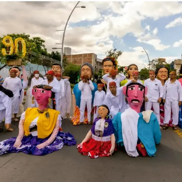 Fotografía de una comparsa o desfile callejero con participantes vestidos de blanco y máscaras o cabezotes gigantes de personajes, posiblemente una manifestación cultural tradicional mexicana.