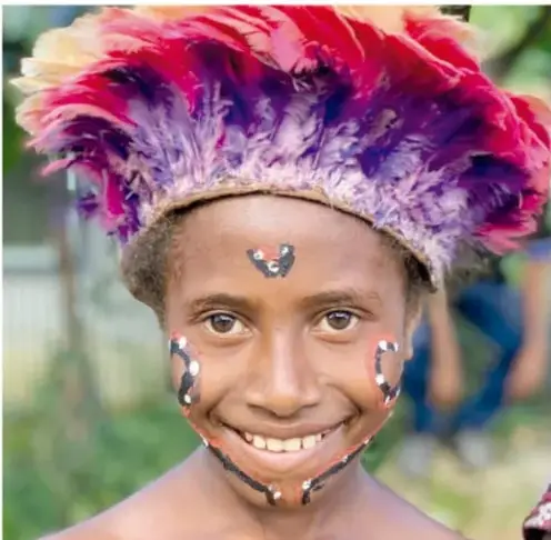 Retrato de un joven sonriente con penacho de plumas de colores (rojo, morado) y pintura decorativa en la cara.