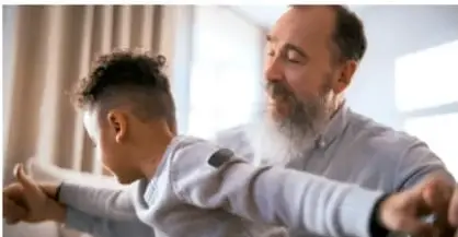 Persona mayor con barba blanca enseñando una práctica corporal o de respiración a un joven dentro de un espacio interior.