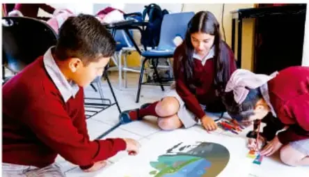 Fotografía de estudiantes de telesecundaria trabajando en equipo en el piso con una gran infografía o mapa sobre contaminación ambiental, utilizando materiales de dibujo.