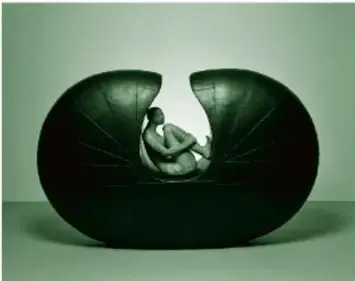 Fotografía de la escultura Trayectos (2001) de Miriam Medrez: forma ovalada de semilla oscura en acero y cerámica, con una mujer de perfil sentada en su interior, rodillas flexionadas, brazos sobre las piernas y cabeza alzada.