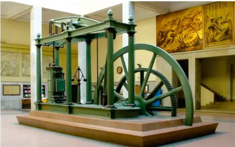 Fotografía de una máquina de vapor histórica (estilo Watt) exhibida en un museo, con gran rueda volante y estructura verde sobre plataforma de madera.