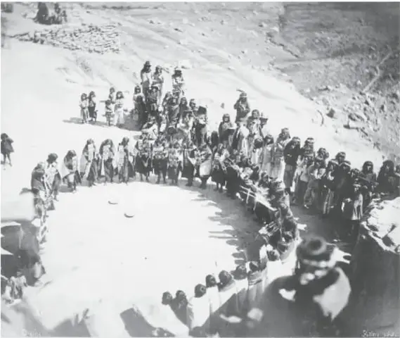 Fotografía en blanco y negro titulada «Danza de la lluvia hopi»: danzante con atuendo tradicional camina frente a un público reunido en terrazas de adobe.