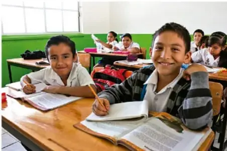 Fotografía de estudiantes de primaria sentados en sus pupitres en un aula, sonriendo y trabajando.