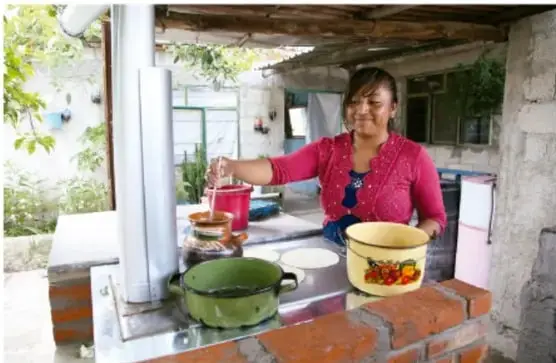 Mujer cocinando en una estufa ecológica con ollas de colores en un espacio semicubierto.