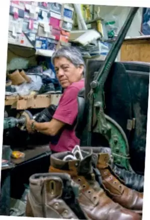 Fotografía de una persona adulta trabajando en un taller de zapatería; se observan estantes con cajas, herramientas y varios pares de botas y zapatos sobre el mostrador.