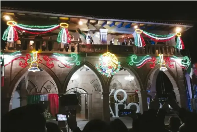 Fotografía nocturna de un edificio con arcos iluminados con luces de colores verde, rojo y blanco, adornado con banderines; al frente hay personas observando, probablemente durante una festividad o conmemoración.