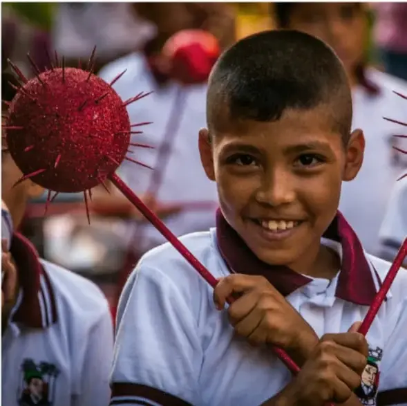 Fotografía de un niño sonriente con uniforme escolar, sosteniendo un objeto decorativo rojo con pinches, en un evento comunitario o escolar; alude a prácticas culturales y artísticas del territorio.