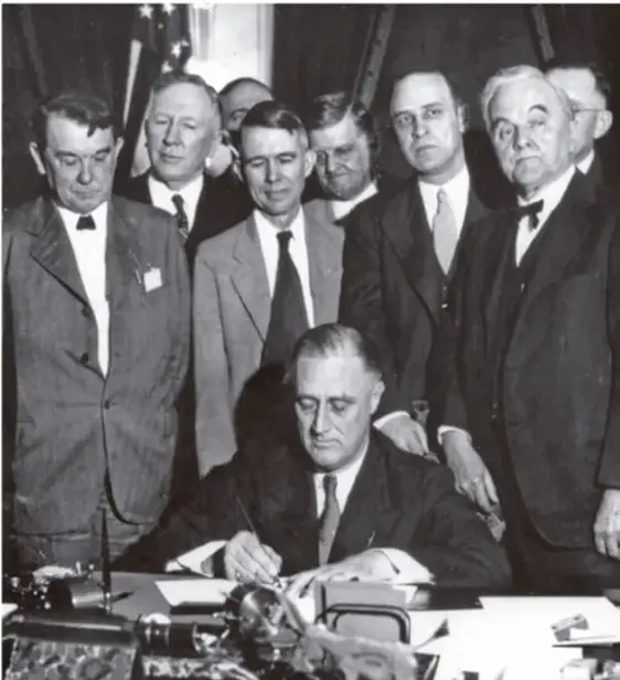 Fotografía en blanco y negro de Franklin D. Roosevelt firmando documentos en su escritorio, rodeado de varios hombres con traje.