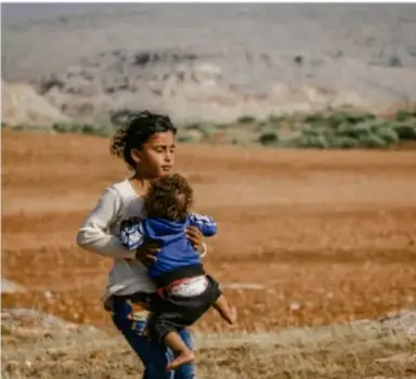 Fotografía de una mujer cargando a un niño pequeño en brazos, caminando por un paisaje árido.