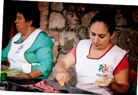 Fotografía de dos mujeres con delantal blanco preparando alimentos en una cocina comunitaria, con pared de piedra al fondo.